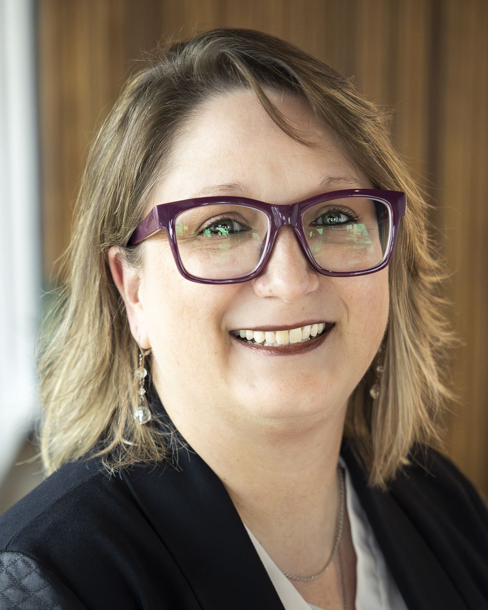 Portrait of a woman wearing large purple eyeglasses, a dark blazer, and a light-colored top, smiling at the camera. The background is softly blurred with warm wood paneling and subtle indoor lighting.