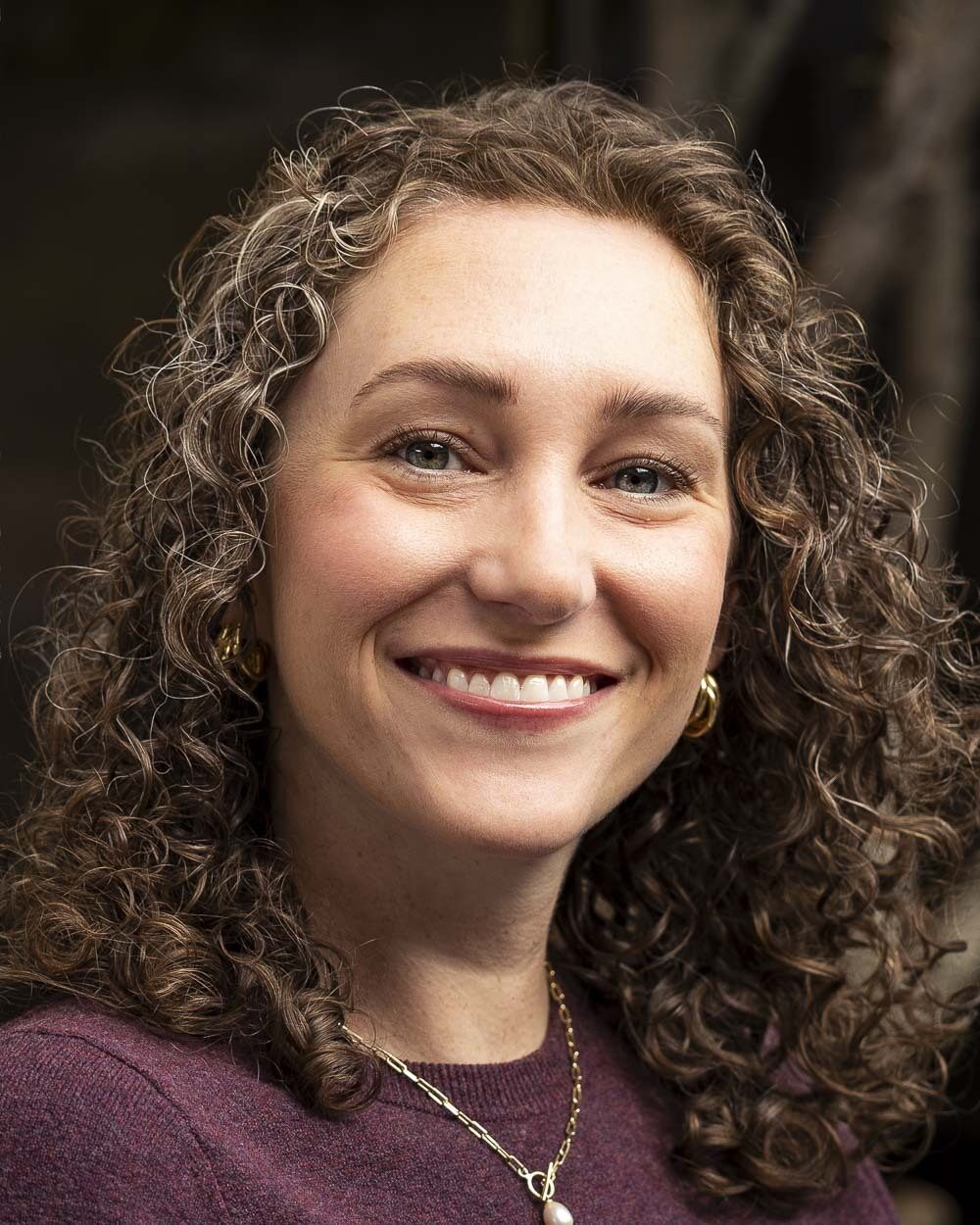 Portrait of Marie Drenon, a woman with shoulder-length curly hair wearing a burgundy sweater, earrings and a necklace, photographed against a softly blurred background.