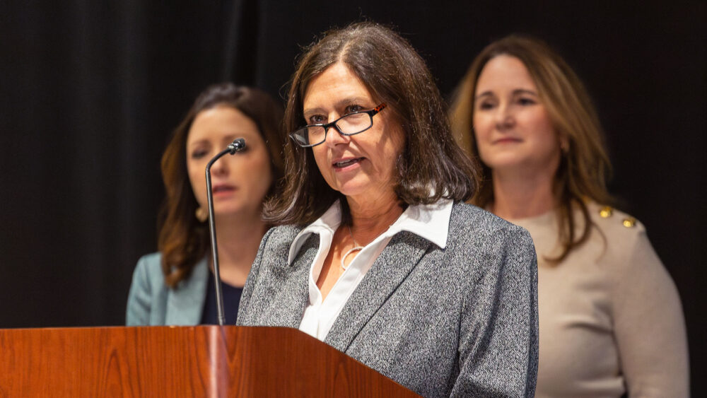A woman wearing reading glasses, a gray blazer and a white collared shirt speaks at a podium. Two women stand behind her.