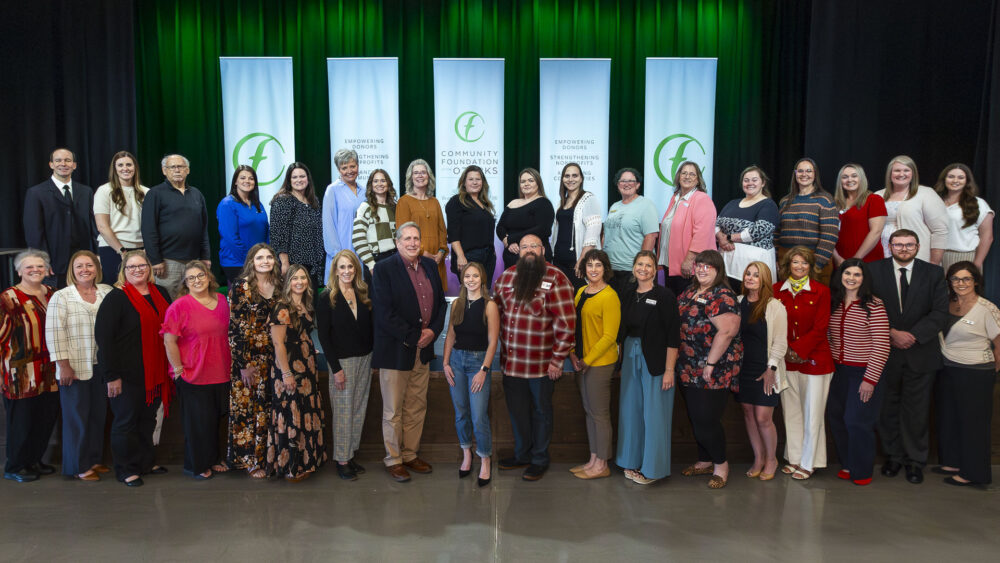 A large group of people standing on a stage for a formal group photo, with five vertical banners reading “Community Foundation of the Ozarks” and messages about empowering donors and strengthening nonprofits behind them, set against a green-lit curtain backdrop.