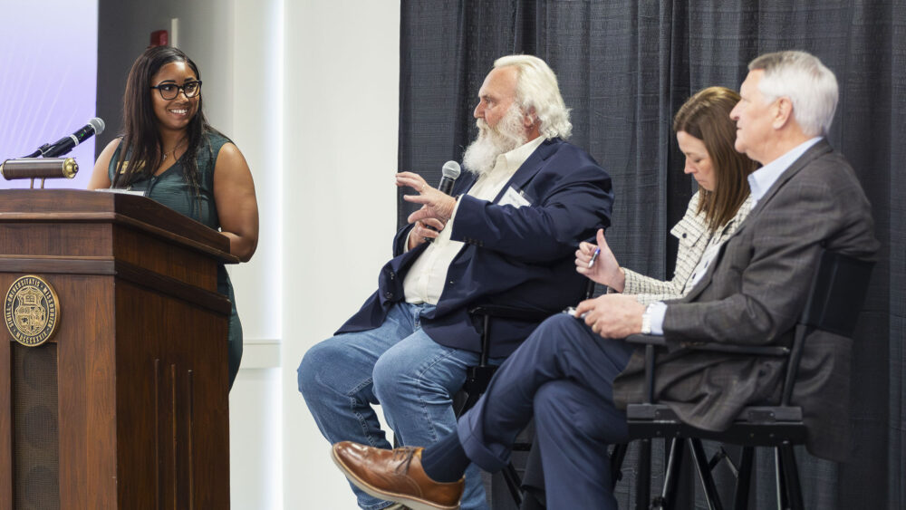 A moderator stands at a podium and addresses a seated panel of four people on stage, with microphones, chairs, and a projection screen visible in a conference setting.