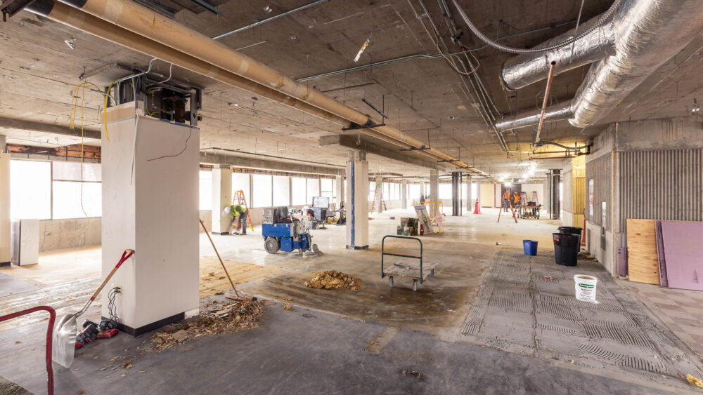 An indoor renovation project site with dusty floors, bare ducts, pipes and wires, and construction workers.