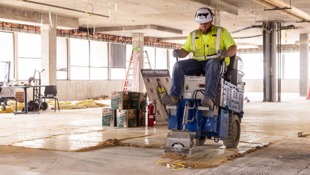 A construction work sits on top of heavy-duty equipment that cleans the floor of old adhesives.