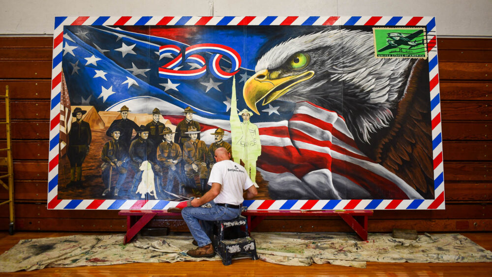 Picture of a man painting a large mural depicting soldiers from World War I, a bald eagle and an American flag.