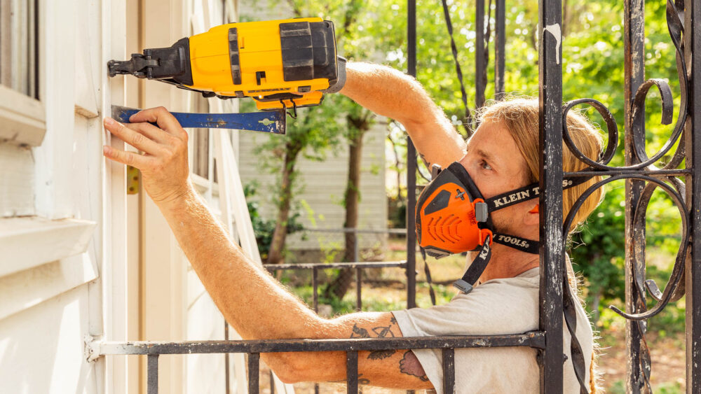A man uses a power tool to work on the exterior of a white house.