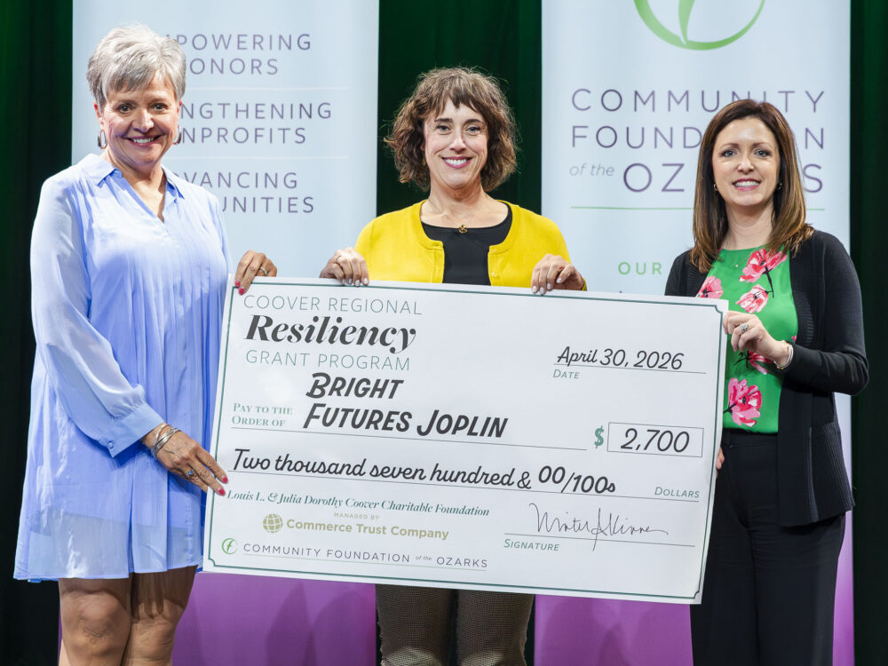 Three people hold a ceremonial $2,700 grant check for Bright Futures Joplin through the Coover Regional Resiliency Grant Program, standing in front of Community Foundation of the Ozarks banners.