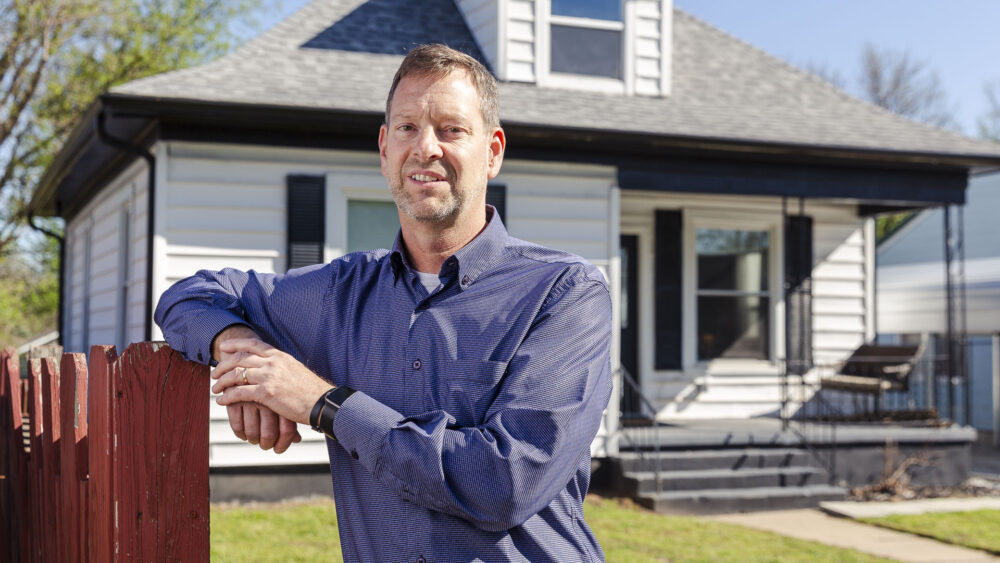 A man in a blue button-down shirt leans against a red wooden fence in front of a white, single-story house with black shutters on a sunny day.