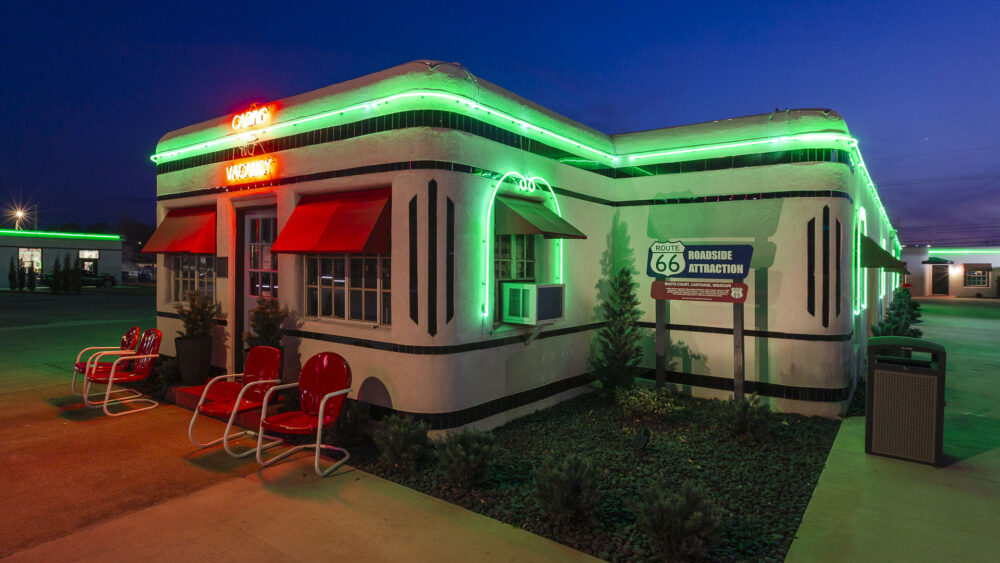 A close-up of the Boots Court motel office at dusk, featuring distinctive Art Deco architecture with white walls and black stripes. Bright green neon tubing outlines the roof and windows, complemented by red metal chairs out front.