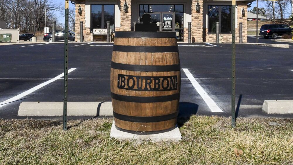 A large wooden barrel with black metal hoops and a sign that reads "BOURBON" sits on a concrete pad in front of a stone-faced building and parking lot.