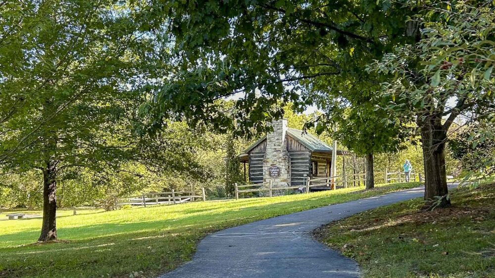 A paved walking path curves through a lush green park toward a small, historic log cabin with a stone chimney, framed by overhanging tree branches.