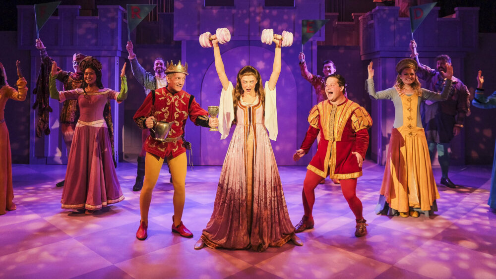 A group of actors in medieval costumes perform on a purple-lit stage. A central woman in a pink gown playfully lifts pink barbell weights while a man in a king’s costume holds a goblet nearby.