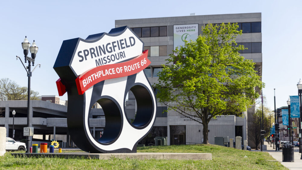 A large, sculptural Route 66 shield sign on a grassy roadside. It reads "Springfield Missouri, Birthplace of Route 66" in bold letters against a backdrop of a modern city office building and a green tree.