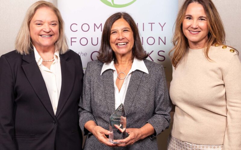 Three women pose in front of a banner. The woman in the center is holding an award made of glass or crystal.