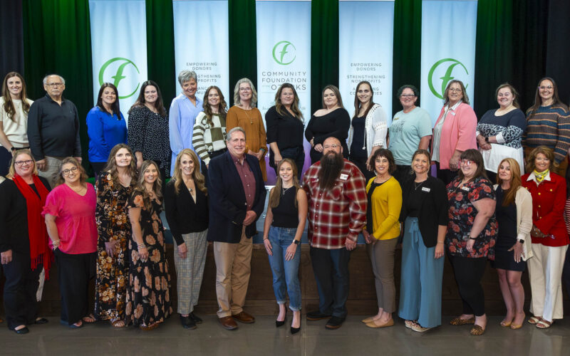 A large group of people standing on a stage for a formal group photo, with five vertical banners reading “Community Foundation of the Ozarks” and messages about empowering donors and strengthening nonprofits behind them, set against a green-lit curtain backdrop.