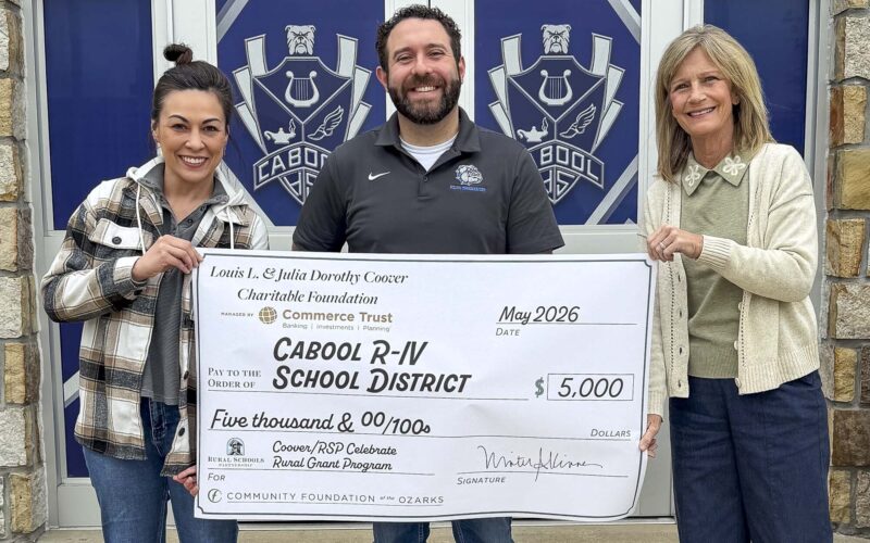Three adults stand in front of a building entrance with panels displaying a bulldog-style school crest, holding a large presentation check. The check, dated May 2026, is made out to “Cabool R‑IV School District” in the amount of $5,000 from the Louis L. & Julia Dorothy Coover Charitable Foundation, managed by Commerce Trust, with “Coover/RSP Celebrate Rural Grant Program” and “Community Foundation of the Ozarks” noted at the bottom.