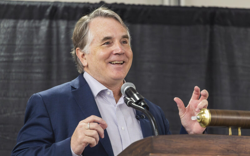 “Close-up of a person standing at a podium and speaking into a microphone, wearing a suit jacket and light-colored shirt, with a dark curtain backdrop.