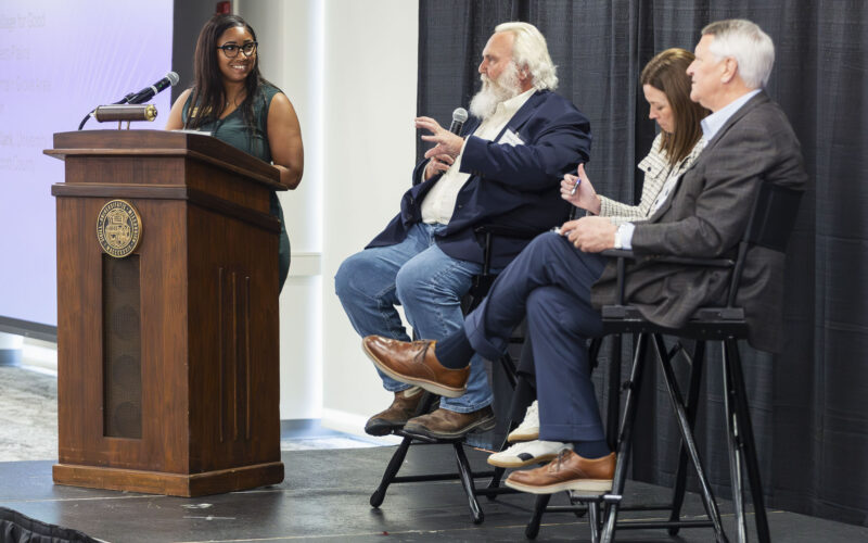 A moderator stands at a podium and addresses a seated panel of four people on stage, with microphones, chairs, and a projection screen visible in a conference setting.