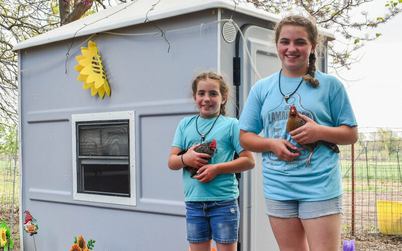 Two girls each hold a chicken as they stand in front of a fiberglass chicken coop.