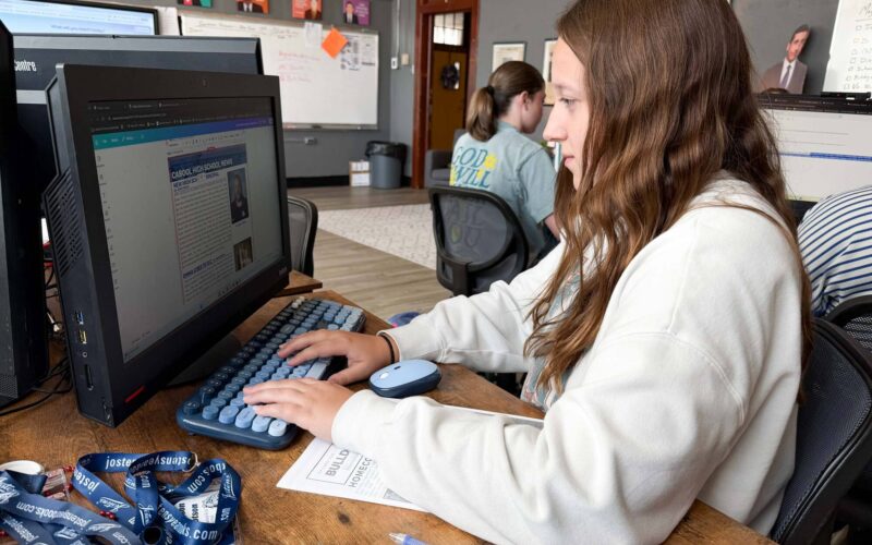 A student works at a desktop computer on a school news publication, with other students and computer workstations visible in the classroom.