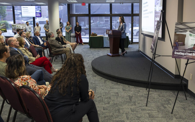 Winter Kinne, a woman wearing a gray blazer, stands at a podium and speaks to a seated audience in a conference room, with slides projected on a screen and campaign display boards for “Creating a Front Door for Philanthropy” visible nearby.