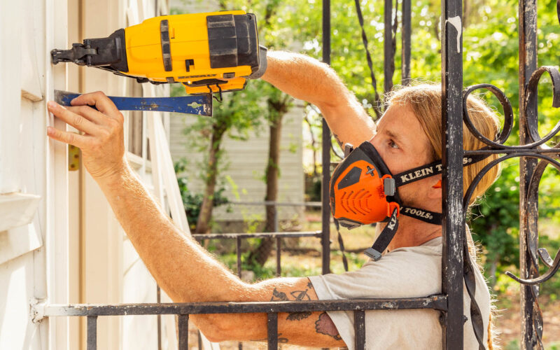 A man uses a power tool to work on the exterior of a white house.