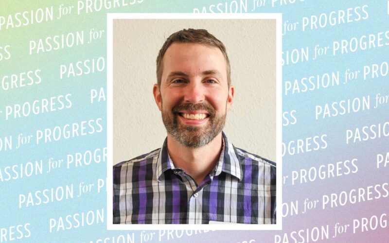 A headshot of Micah Titterington, a man with a short beard smiling at the camera. He is wearing a purple and grey plaid button-down shirt against a background featuring the repeating phrase "PASSION for PROGRESS."