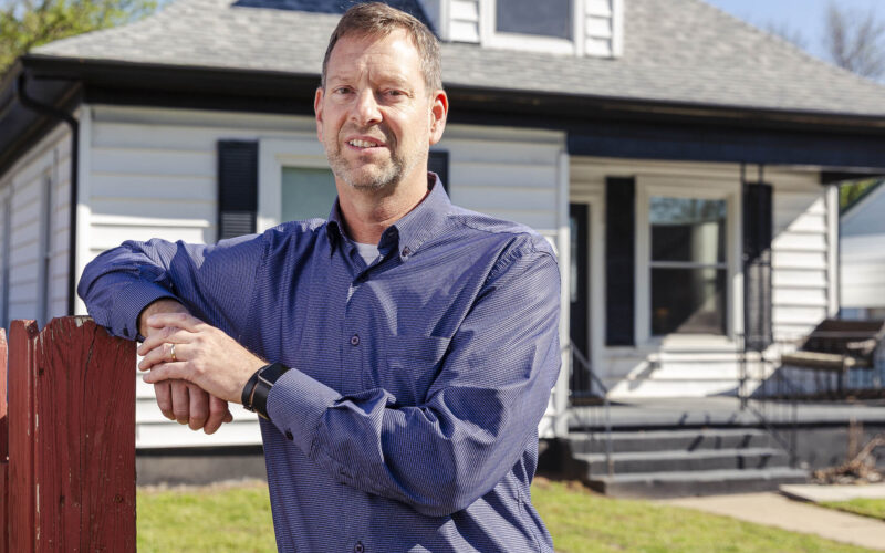 A man in a blue button-down shirt leans against a red wooden fence in front of a white, single-story house with black shutters on a sunny day.