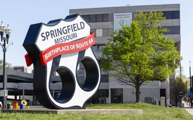 A large, sculptural Route 66 shield sign on a grassy roadside. It reads "Springfield Missouri, Birthplace of Route 66" in bold letters against a backdrop of a modern city office building and a green tree.