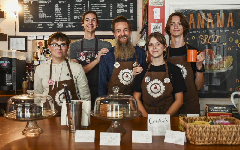 Five smiling employees of the Firehouse Coffee Shop stand behind a wooden counter wearing brown aprons. Trays of pastries in glass domes are in the foreground.