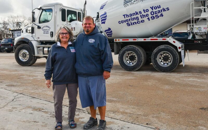 A woman and a man pose in front of a white concrete truck. Printing on the truck reads Thanks to You covering the Ozarks Since 1954.