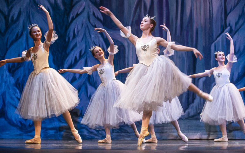 Ballet dancers in white tutus and tiaras perform onstage during a production of The Nutcracker, posing en pointe against a blue winter-themed backdrop.