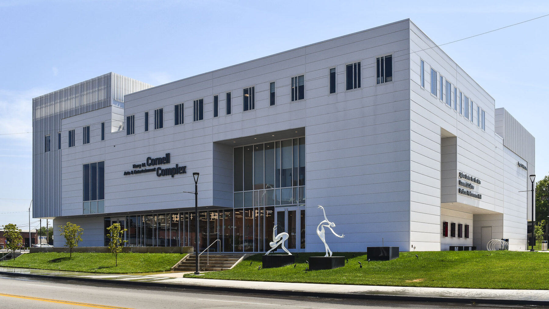 The exterior of the Harry M. Cornell Arts & Entertainment Complex, a modern white three-story building with large glass windows and two white abstract dancer sculptures on the front lawn.