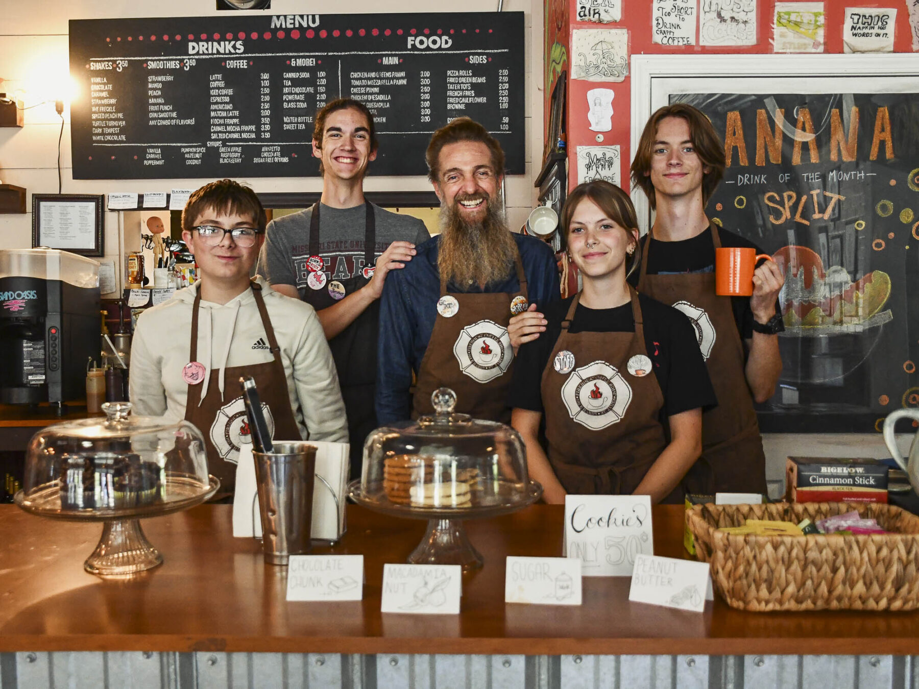 Five smiling employees of the Firehouse Coffee Shop stand behind a wooden counter wearing brown aprons. Trays of pastries in glass domes are in the foreground.