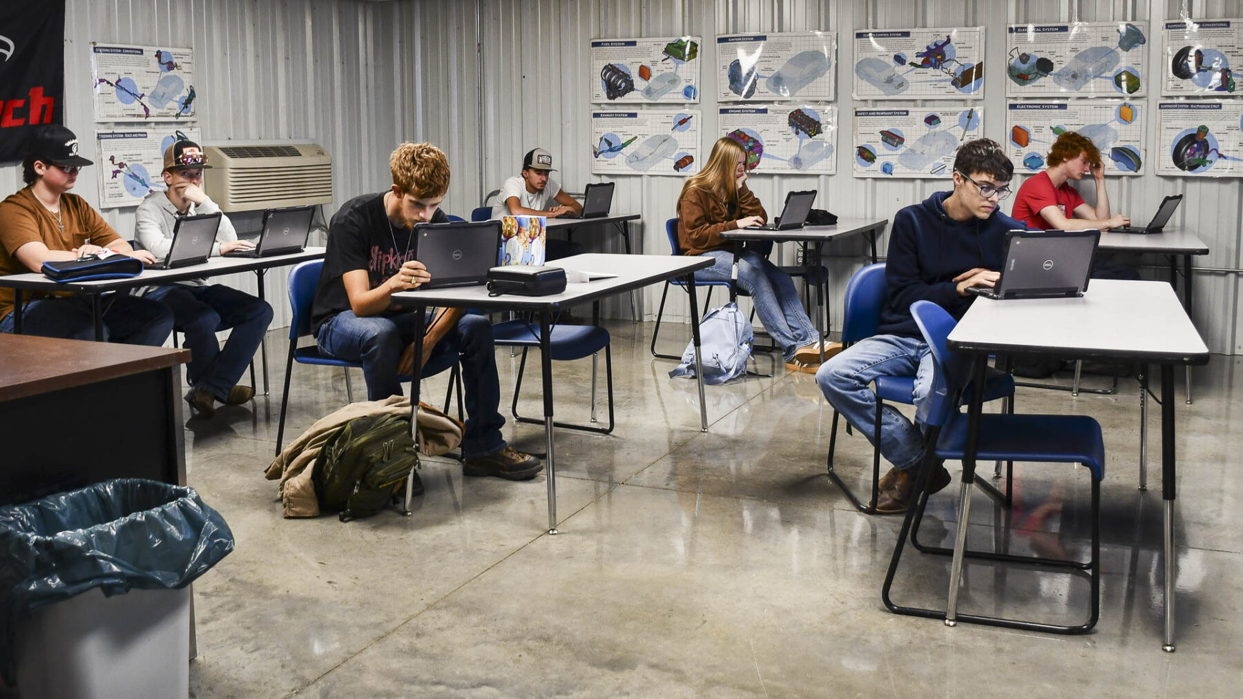 Students work individually on laptops at tables in a classroom with automotive system diagrams displayed on the walls.