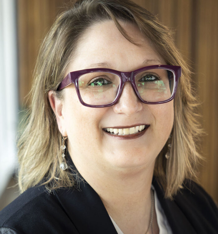 Portrait of a woman wearing large purple eyeglasses, a dark blazer, and a light-colored top, smiling at the camera. The background is softly blurred with warm wood paneling and subtle indoor lighting.