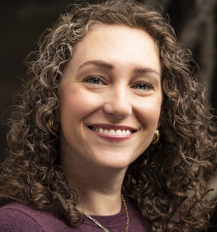 Portrait of Marie Drenon, a woman with shoulder-length curly hair wearing a burgundy sweater, earrings and a necklace, photographed against a softly blurred background.