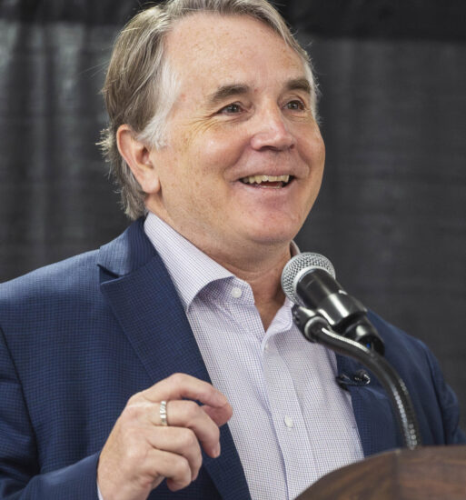 “Close-up of a person standing at a podium and speaking into a microphone, wearing a suit jacket and light-colored shirt, with a dark curtain backdrop.