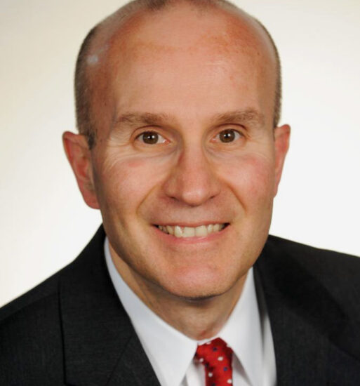Portrait of Hank Molinengo, a man wearing a dark suit, white shirt and red patterned tie against a light background.