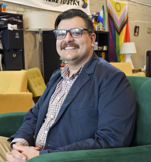 Portrait of Aaron Kitchens, a man wearing glasses and a blazer seated in a community center lounge, with Pride flags and a banner reading “We Are Here to Stay” displayed in the background.