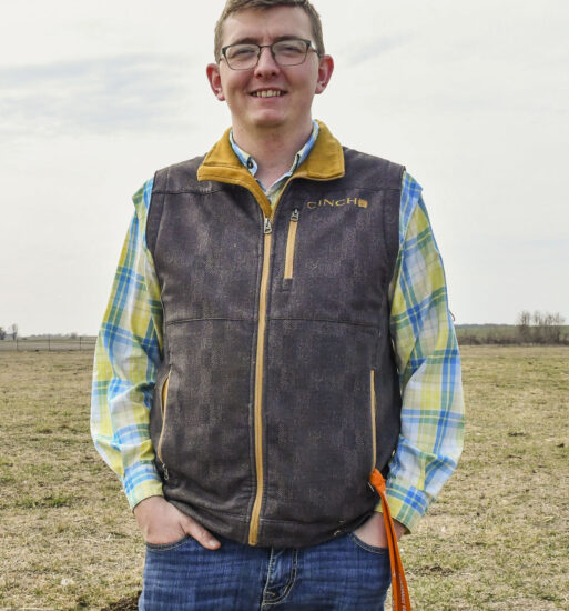 Portrait of Michael Lee, a man wearing glasses, a plaid shirt and a zip-up vest, standing in a pasture.