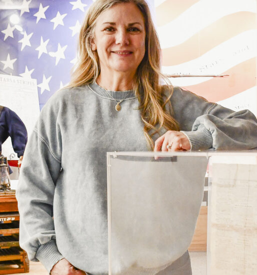 Portrait of Laura Dumey, a woman standing beside a display case in a museum-like setting, with an American flag mural and historical artifacts visible in the background.