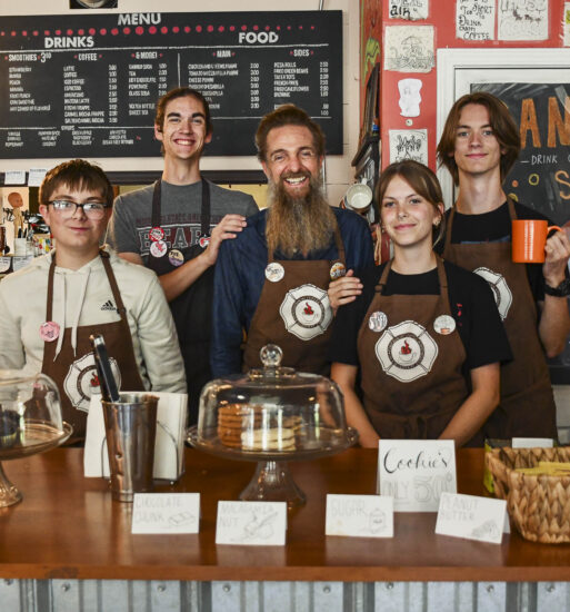 Five smiling employees of the Firehouse Coffee Shop stand behind a wooden counter wearing brown aprons. Trays of pastries in glass domes are in the foreground.