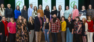 A large group of people standing on a stage for a formal group photo, with five vertical banners reading “Community Foundation of the Ozarks” and messages about empowering donors and strengthening nonprofits behind them, set against a green-lit curtain backdrop.