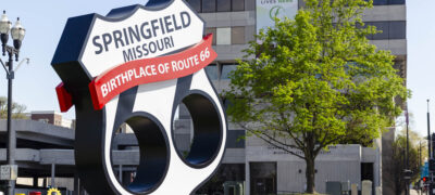 A large, sculptural Route 66 shield sign on a grassy roadside. It reads "Springfield Missouri, Birthplace of Route 66" in bold letters against a backdrop of a modern city office building and a green tree.