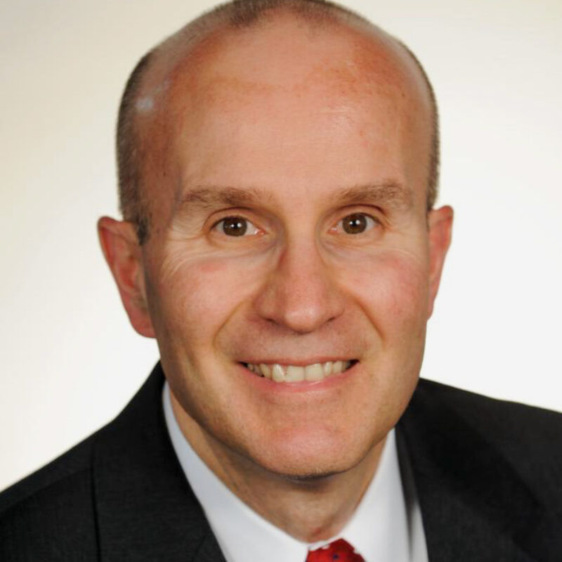 Portrait of Hank Molinengo, a man wearing a dark suit, white shirt and red patterned tie against a light background.