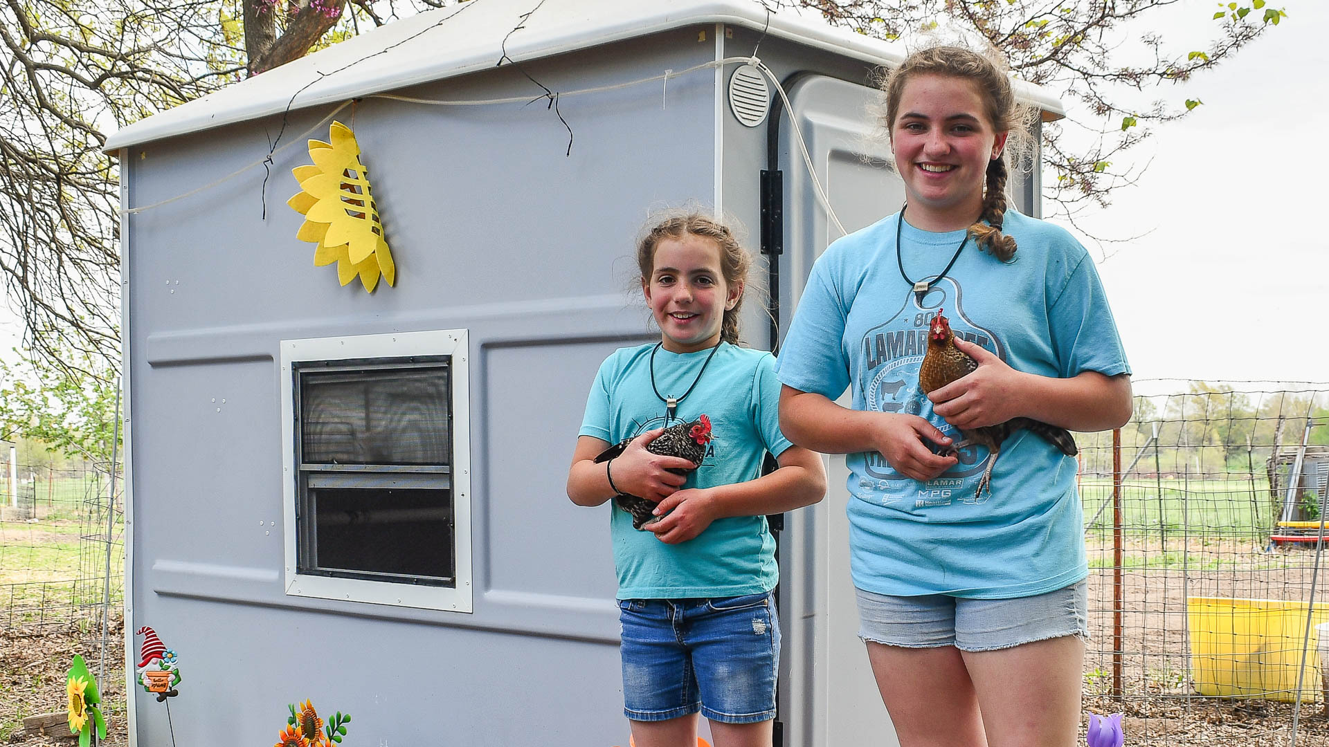 Two girls each hold a chicken as they stand in front of a fiberglass chicken coop.